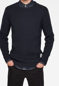 Granatowy sweter dzianinowy o żeberkowej strukturze z okrągłym dekoltem i długimi rękawami, nałożony na dżinsową koszulę, w połączeniu z czarnymi spodniami.