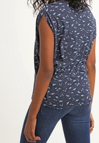 Navyblå ärmlös skjorta med ljusblå fågelmönster, mjukt tyg, rund halsringning och avslappnad passform. Matchas med mörka denimjeans.