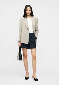 Jeune femme portant un blazer beige rayé, un haut blanc, un short bleu marine et des escarpins noirs, tenant un petit sac à main noir.