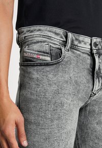 Jeans negros desgastados con corte ajustado, que presentan un bolsillo frontal con una etiqueta en contraste, textura suave y costuras de denim tradicionales.