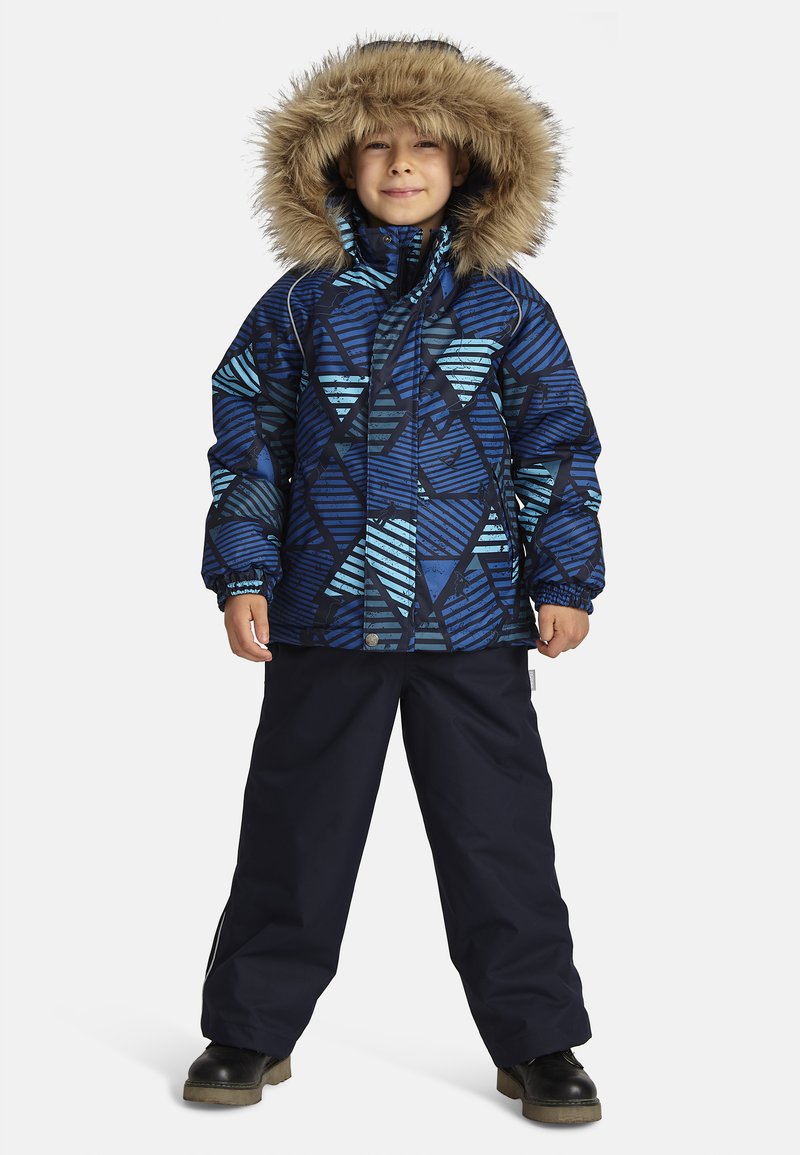 Blå pufferjacka med geometriskt mönster och avtagbar fuskpäls huva, parat med mörka byxor och svarta stövlar, stående på en vit bakgrund.