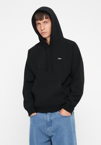 Sort hættebluse med lomme foran og "OBEY" logo i hvid. Stoffet ser blødt ud med en afslappet pasform, som suppleres af blå denimbukser.