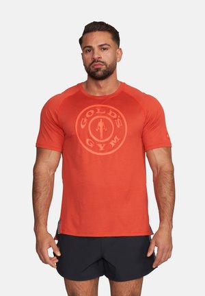 Mężczyzna z brodą i krótkimi włosami, ubrany w czerwoną koszulkę Gold's Gym i czarne spodenki, stoi na białym tle.