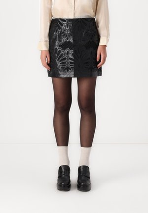 Vrouw draagt een zwarte leren minirok met bloempatronen, zwarte transparante panty's, witte sokken, zwarte loafers en een crèmekleurige blouse.