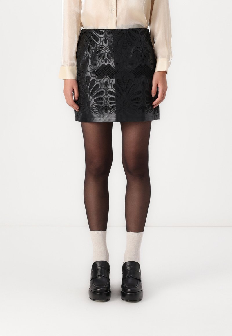 Femme portant une mini-jupe en cuir noire à motifs floraux, des collants noirs transparents, des chaussettes blanches, des mocassins noirs et un chemisier crème.