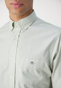 Light green button-up shirt with a collar, featuring a pocket and a small embroidered logo on the chest. Smooth fabric texture.