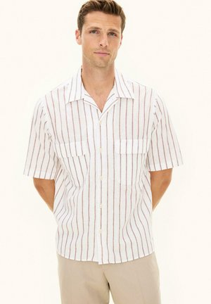 Mężczyzna w białej koszuli z krótkim rękawem i beżowymi pionowymi paskami oraz beżowych spodniach, stojący z rękami za plecami.