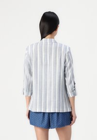 Blazer rayé en tissu léger avec des rayures verticales bleues et blanches, manches trois-quarts et col cranté. Porté par-dessus un short bleu à pois.