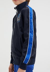 Lapsi, joka on pukeutunut tummansiniiseen vetoketjulliseen takkiin, jonka hihoissa on sinivalkoiset raidat, ja rentoihin sinisiin housuihin, yksinkertaista taustaa vasten.