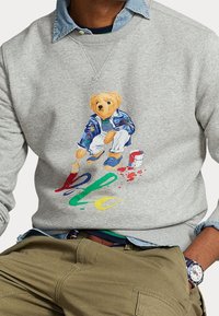 Grå sweatshirt med en illustrerad björn i en färgglad outfit, som håller en pensel med färgglada färgpluppar och "Hej" i livfulla färger.