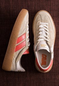 Paire de baskets en daim beige avec semelles en gomme et bandes rouges, lacets blancs et logo « CAVAL » sur la languette et la semelle intérieure, sur une surface texturée foncée.