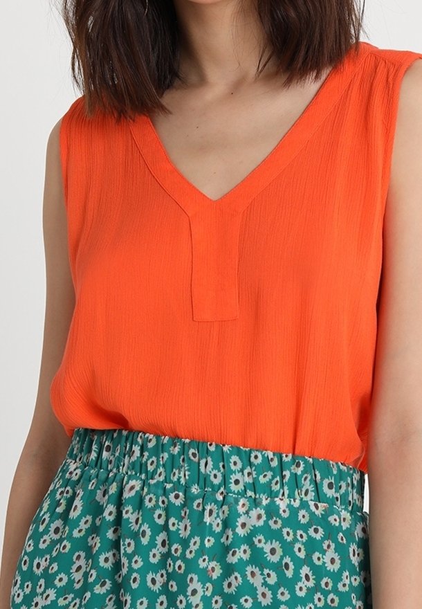 Orange ærmeløs bluse med V-hals og en tekstureret finish, parret med et grønt blomsterprintet skørt med elastik i taljen.