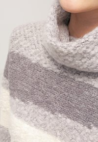 Světle šedý a krémový pruhovaný svetr s vysokým, texturovaným límcem. Materiál vypadá měkce, má hebký vzhled a útulnou texturu.