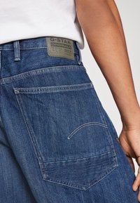 Primer plano de una persona que lleva puestos jeans de mezclilla G-Star RAW azules con el bolsillo trasero visible y la etiqueta de la marca, combinados con una camisa blanca.