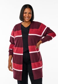 Leende kvinna med axellångt svart hår bär en lång vinröd cardigan med röda och rosa ränder över en svart topp och svarta byxor.