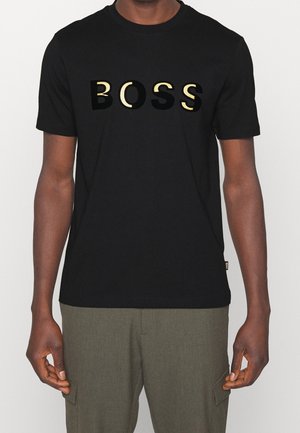 Mann trägt ein schwarzes T-Shirt mit kurzen Ärmeln und großem gold-schwarzem "BOSS"-Schriftzug sowie olivgrüne Hose, steht vor einfarbigem Hintergrund.