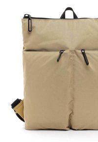 Mochila de nylon beige con cierre superior, dos bolsillos frontales con tiradores de cremallera negros y una correa ajustable negra. Textura suave.