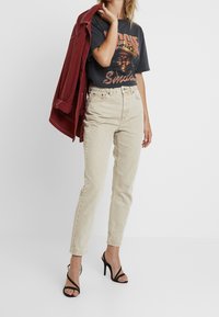 Jeans de tiro alto beige combinados con una camiseta gráfica negra que presenta texto en naranja y una chaqueta burdeos. Se llevan tacones altos negros con tiras.