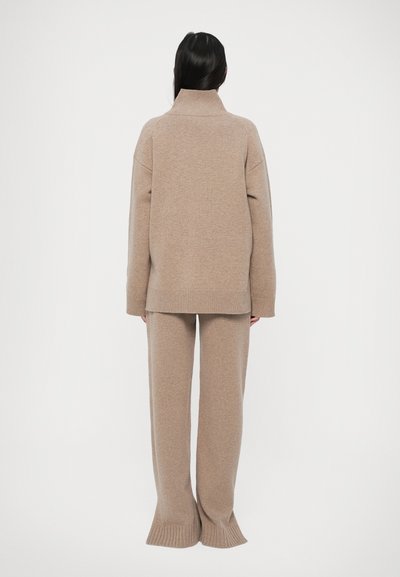 Persoon met lang, donker haar die een beige gebreide trui met hoge hals en bijpassende wijde broek draagt, staand met de rug naar de camera op een witte achtergrond.