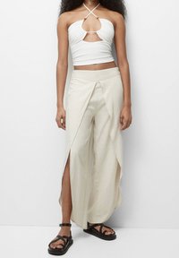 Haut halter blanc avec un design découpé, associé à un pantalon large beige comportant des fentes latérales et un tissu lisse et texturé. Sandales noires.