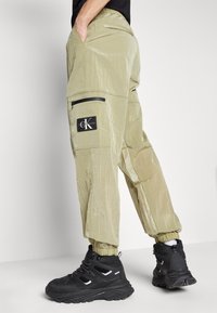 Svijetlo maslinaste cargo hlače s sjajnom, naboranom teksturom, elastičnim manžetama, bočnim džepovima i crnom Calvin Klein logom. Nosene s crnim tenisicama.