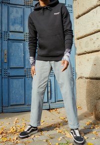 Felpa nera con cappuccio e tasca frontale, pantaloni a sigaretta blu chiaro e sneaker bianche e nere. In piedi di fronte a una porta blu con graffiti.