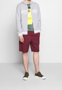 Felpa grigia con zip e logo bianco, t-shirt verde a strati sotto, pantaloni corti bordeaux e sneakers verde oliva con suola bianca.