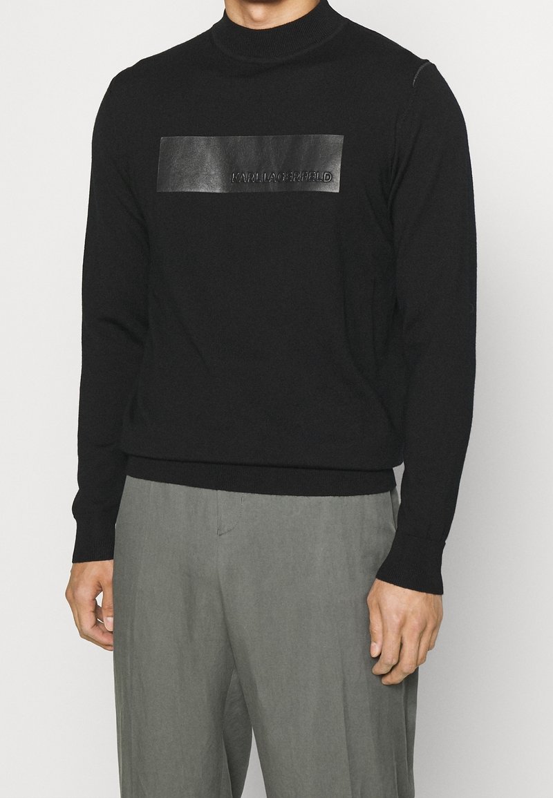 Svart stickad tröja med hög krage, med en texturerad svart läderlapp med präglad logotyp. Figurpassad design med ribbade ärmslut och nederkant.