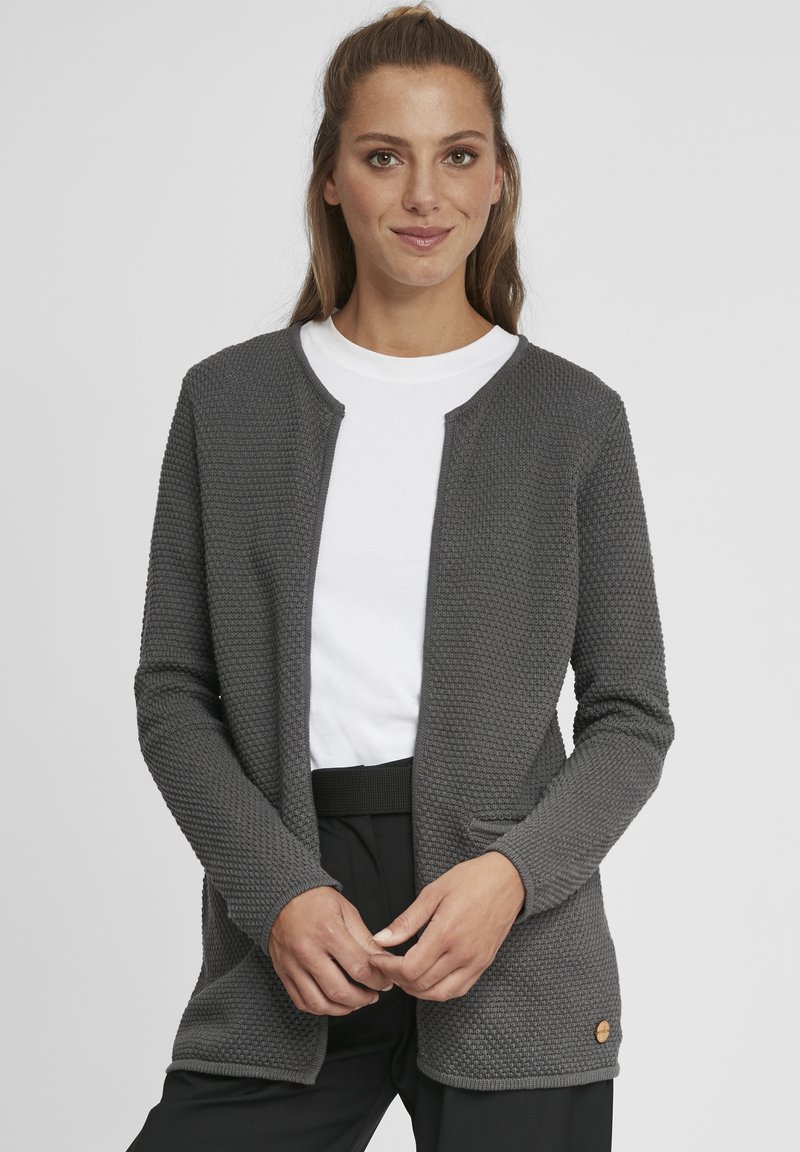 Gray textured cardigan with a rounded neckline, open front, side pockets, and a wooden accent near the hem, worn over a white t-shirt.