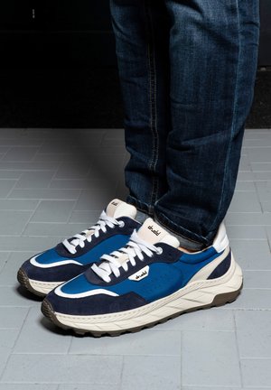 Persoon die blauwe en witte sneakers draagt met witte veters en donkerblauwe spijkerbroek, staand op een lichtgrijze tegelvloer.