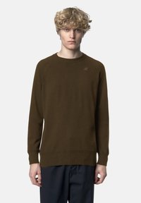 Maglione marrone lavorato a maglia con scollo rotondo e polsini a coste. Presenta un dettaglio discreto del logo sul petto. Indossato con pantaloni scuri.
