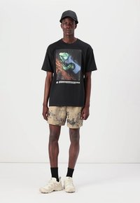 Sort kortærmet t-shirt med grafisk print på fronten, parret med brune shorts med et tekstureret mønster. Footwear er cremefarvet.
