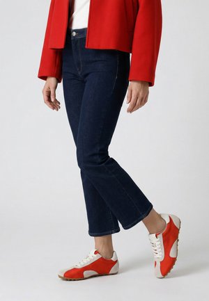 Persoon die donkerblauwe jeans, een rood jasje en witte en rode sneakers draagt, staat tegen een effen achtergrond.