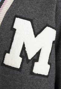 Grand écusson blanc en forme de lettre M avec une bordure noire cousue sur un tissu tricoté gris foncé.