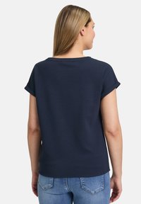 Mørkeblå kortærmet top med rullede opkraver, rund halsudskæring og struktureret stof, parret med lyseblå jeans. Vist fra bagfra.