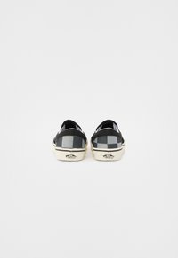 Zapatos de slip-on a cuadros negros y grises con suela de goma y acentos blancos, que presentan un logotipo en el talón y una parte superior de tela suave.
