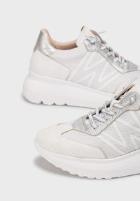 Baskets blanches avec un matériau texturé ressemblant à de la peau, des accents argentés et des panneaux en maille. Semelle épaisse blanche et lacets plats. Présence de lignes de couture décoratives.