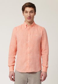 Chemise à manches longues orange clair avec un tissu texturé ; possède un col boutonné et un petit logo blanc sur la poitrine.