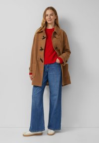 Brązowy wełniany płaszcz z zapięciem na bigle, czerwony sweter w warkocze, niebieskie spodnie typu wide-leg i białe sneakersy z brązowymi podeszwami.
