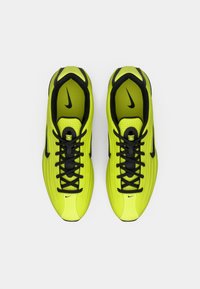 Heledad kollase-rohelised Nike jooksutossud mustade paelte ja swooshi logodega, vaadatuna ülhäält valge tausta pealt.