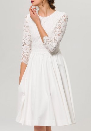 Robe blanche avec un corsage en dentelle à manches trois-quarts, taille froncée, et une jupe évasée avec poches. Texture de tissu lisse.
