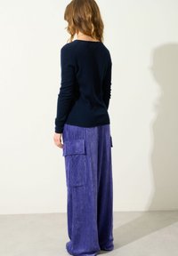 Marineblauer Strickpullover mit langen Ärmeln, kombiniert mit lockeren lila Cordhosen, die zwei große Seitentaschen haben.