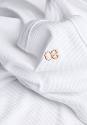 Witte ribstof met een zachte textuur heeft een klein lichtbruin geborduurd logo "oe." De stof valt in plooien, wat duidt op flexibiliteit.