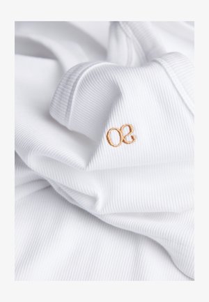 Witte ribstof met een zachte textuur heeft een klein lichtbruin geborduurd logo "oe." De stof valt in plooien, wat duidt op flexibiliteit.