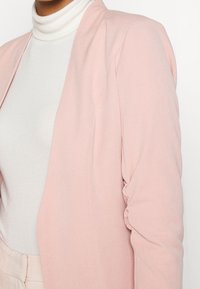 Růžový blazer v odstínu blush s hladkou texturou, přiléhavým střihem, nařasenými rukávy a otevřeným předním dílem, doplněný bílým rolákem pod ním.