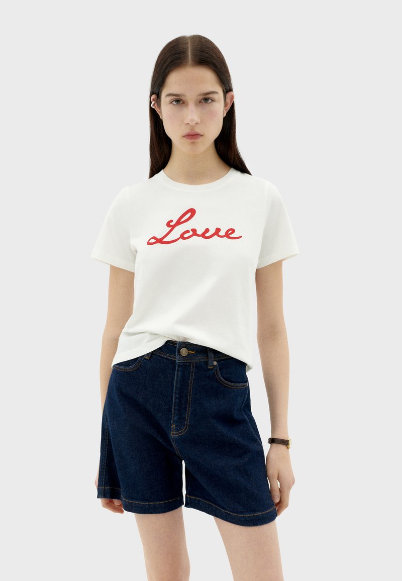 Camiseta de algodón blanca con texto en cursiva rojo que dice "Amor", combinada con pantalones cortos de mezclilla oscura, mostrando un estilo casual y moderno.