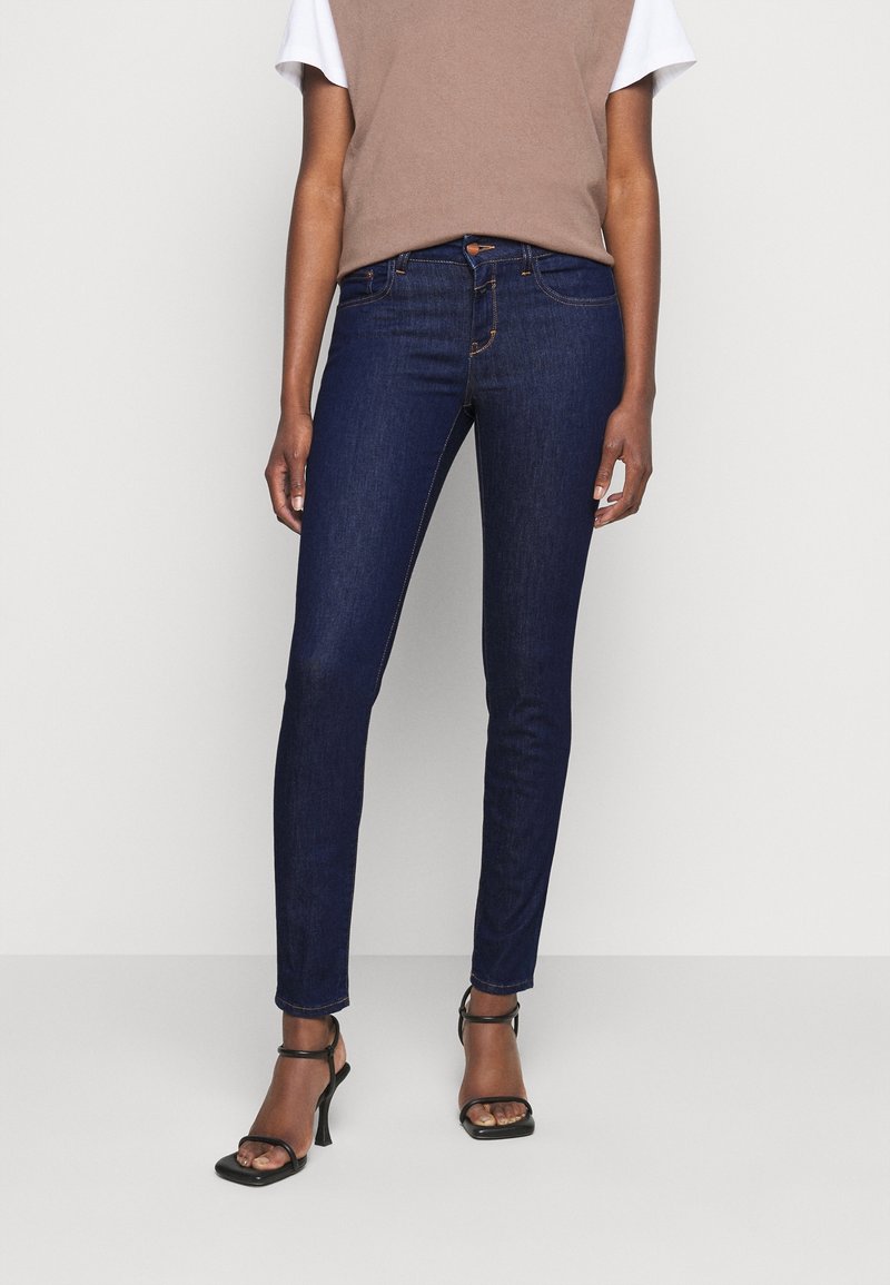 Femme porte un jean skinny bleu foncé, un haut taupe par-dessus un t-shirt blanc et des sandales à talons noires, debout devant un fond clair uni.