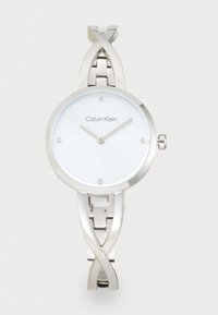 Montre en acier inoxydable argenté avec un cadran rond blanc, des aiguilles fines, quatre petits cristaux et un design de bracelet tordu. Logo Calvin Klein affiché.