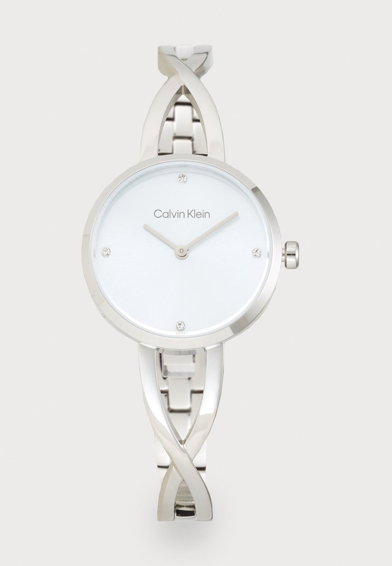 Montre en acier inoxydable argenté avec un cadran rond blanc, des aiguilles fines, quatre petits cristaux et un design de bracelet tordu. Logo Calvin Klein affiché.