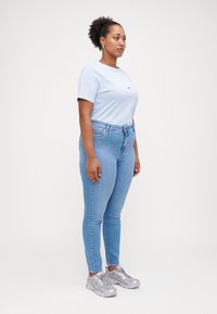 Mujer de pie de perfil, vistiendo una camiseta ajustada de color azul claro, jeans skinny de color azul claro, zapatillas plateadas, con el cabello en un moño rizado.